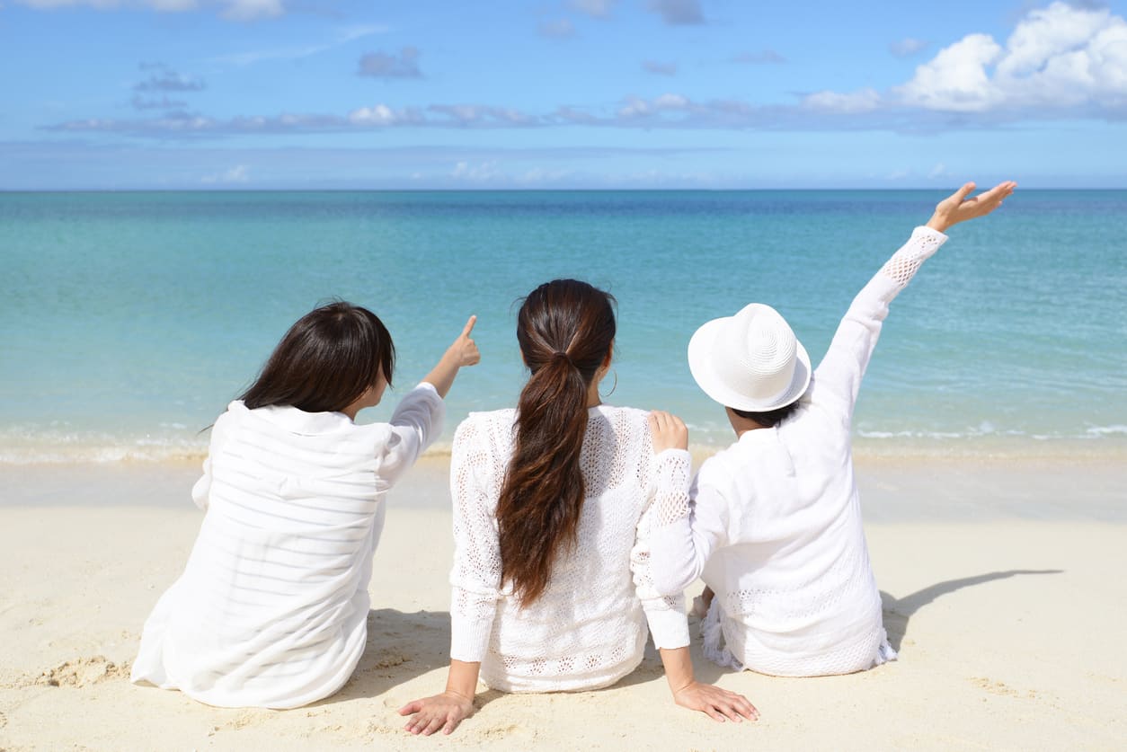 Three friends sitting on a sandy beach, facing the calm blue sea under a clear sky, capturing the peaceful spirit of Marine Day.