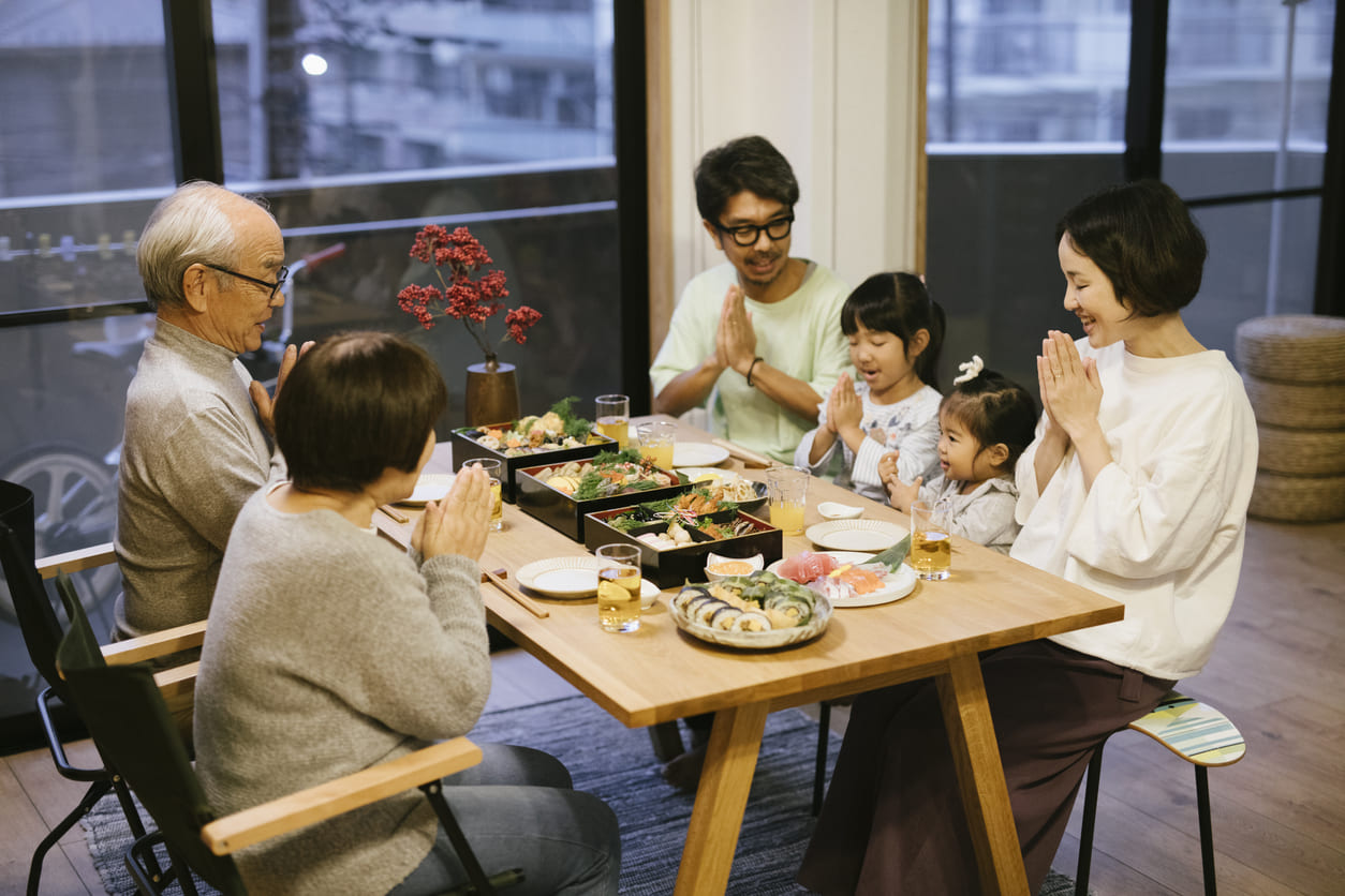 A multi-generational Japanese family seated at a table, sharing a joyful New Year's meal with traditional osechi ryōri and offering a prayer before eating.