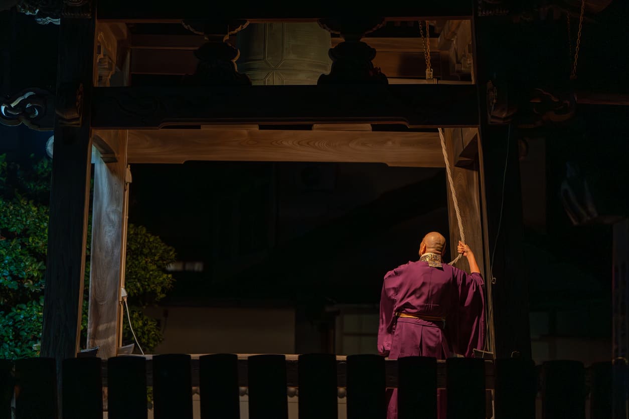 A Buddhist monk in traditional robes ringing a large temple bell at night, capturing the solemn beauty of the joya no kane ritual on New Year's Eve.