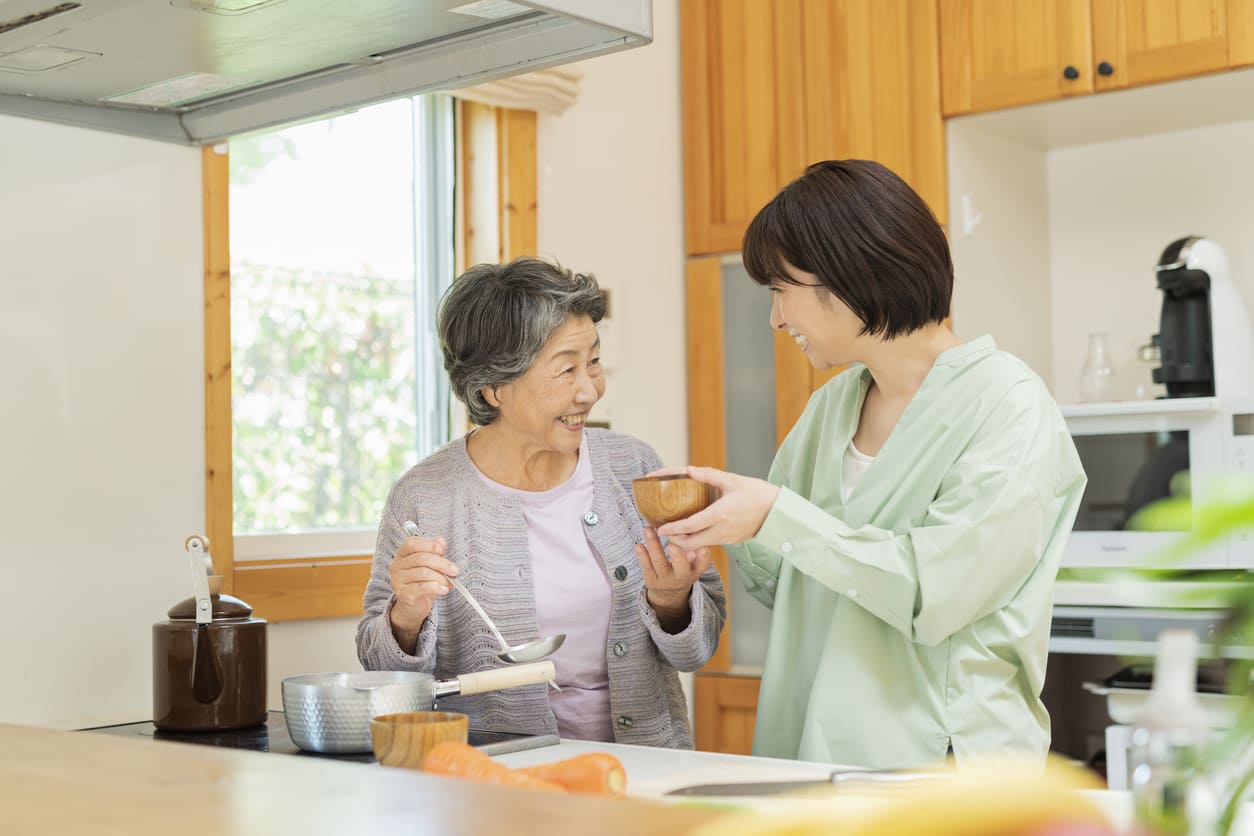 A smiling elderly woman and her younger family member sharing a warm conversation while cooking together in a cozy kitchen.