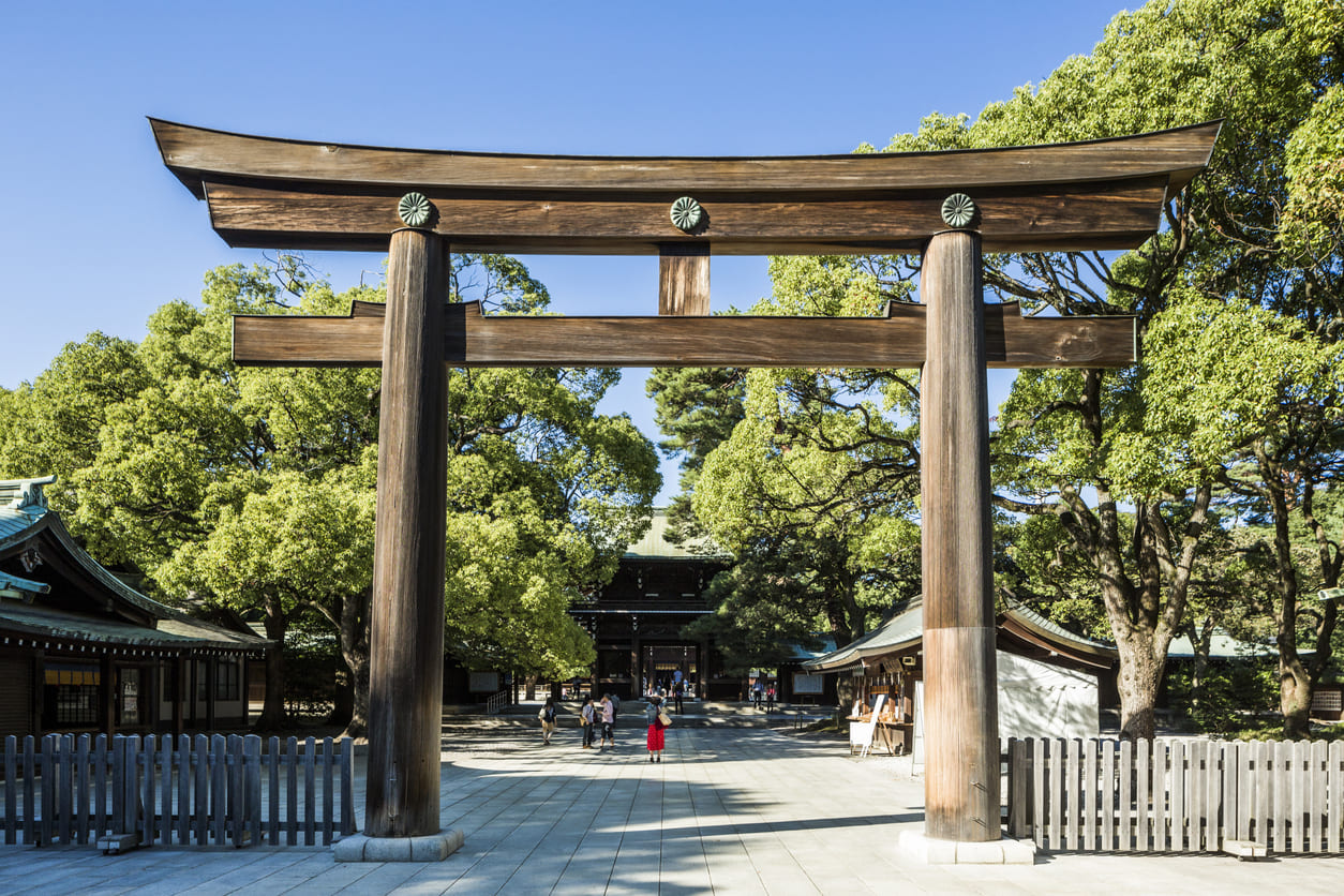 A large wooden torii gate stands at the entrance of Meiji Jingu Shrine, surrounded by lush greenery and a few visitors strolling peacefully beneath it.