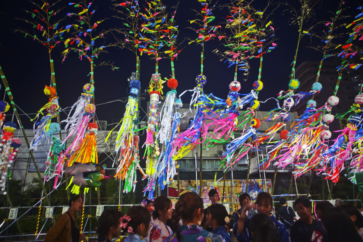 A lively night scene with people in traditional attire gathered beneath tall bamboo poles adorned with bright, flowing Tanabata streamers and paper decorations.