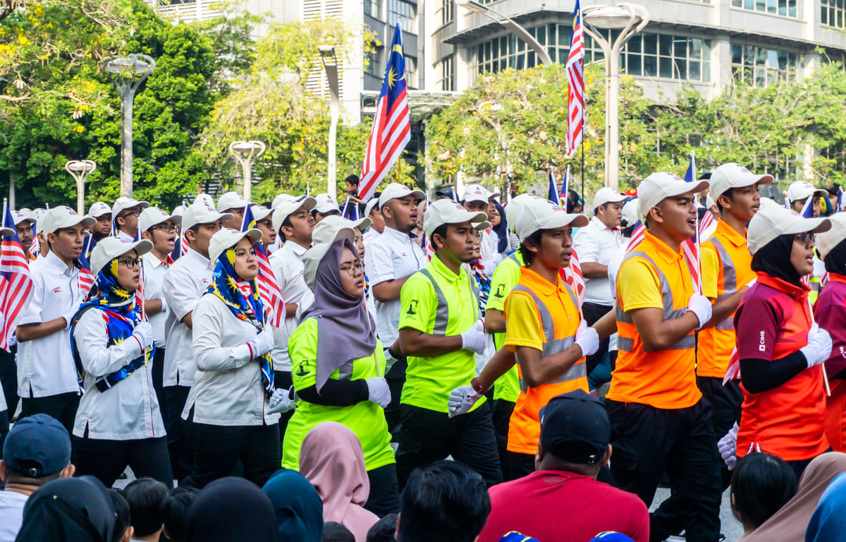 Malaysian workers in uniforms and safety vests marching with flags during a Labour Day parade, watched by a supportive crowd.