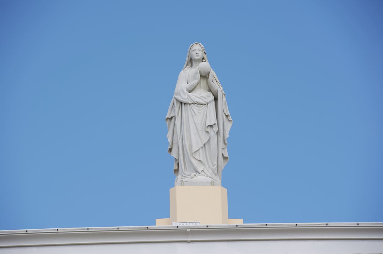 A graceful white statue of the Virgin Mary stands atop Marie Reine de la Paix, set against a clear blue sky, embodying peace and devotion.