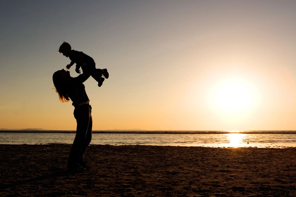A silhouette of a mother joyfully lifting her child by the beach during a golden sunset.