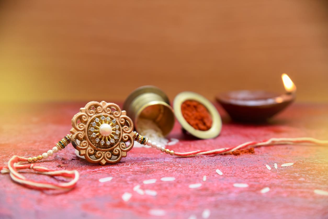 A decorative rakhi rests on a vibrant surface beside kumkum, rice, and a lit oil lamp, capturing the essence of Raksha Bandhan rituals.