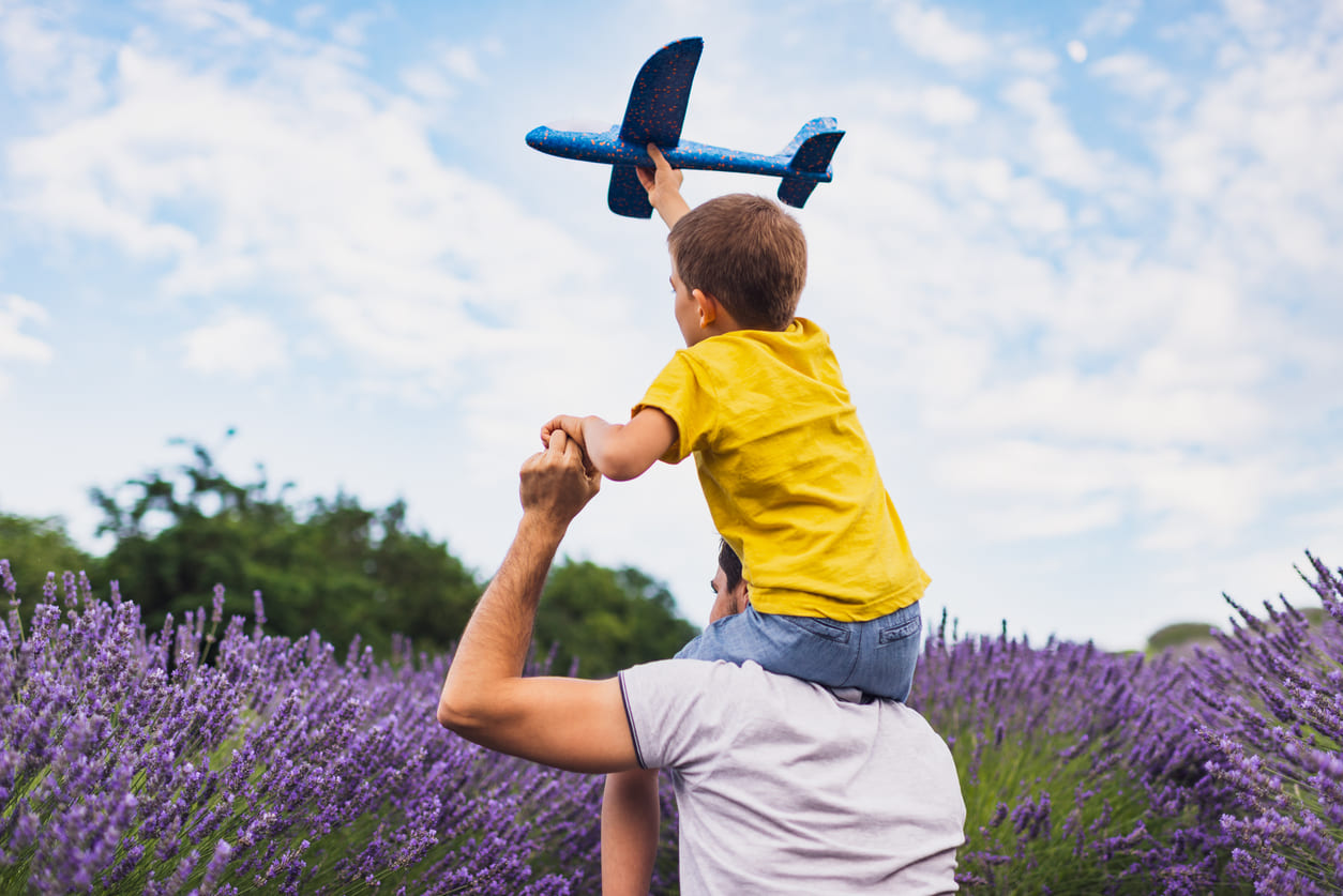 A child sits on their father's shoulders, joyfully flying a toy airplane in a picturesque lavender field.