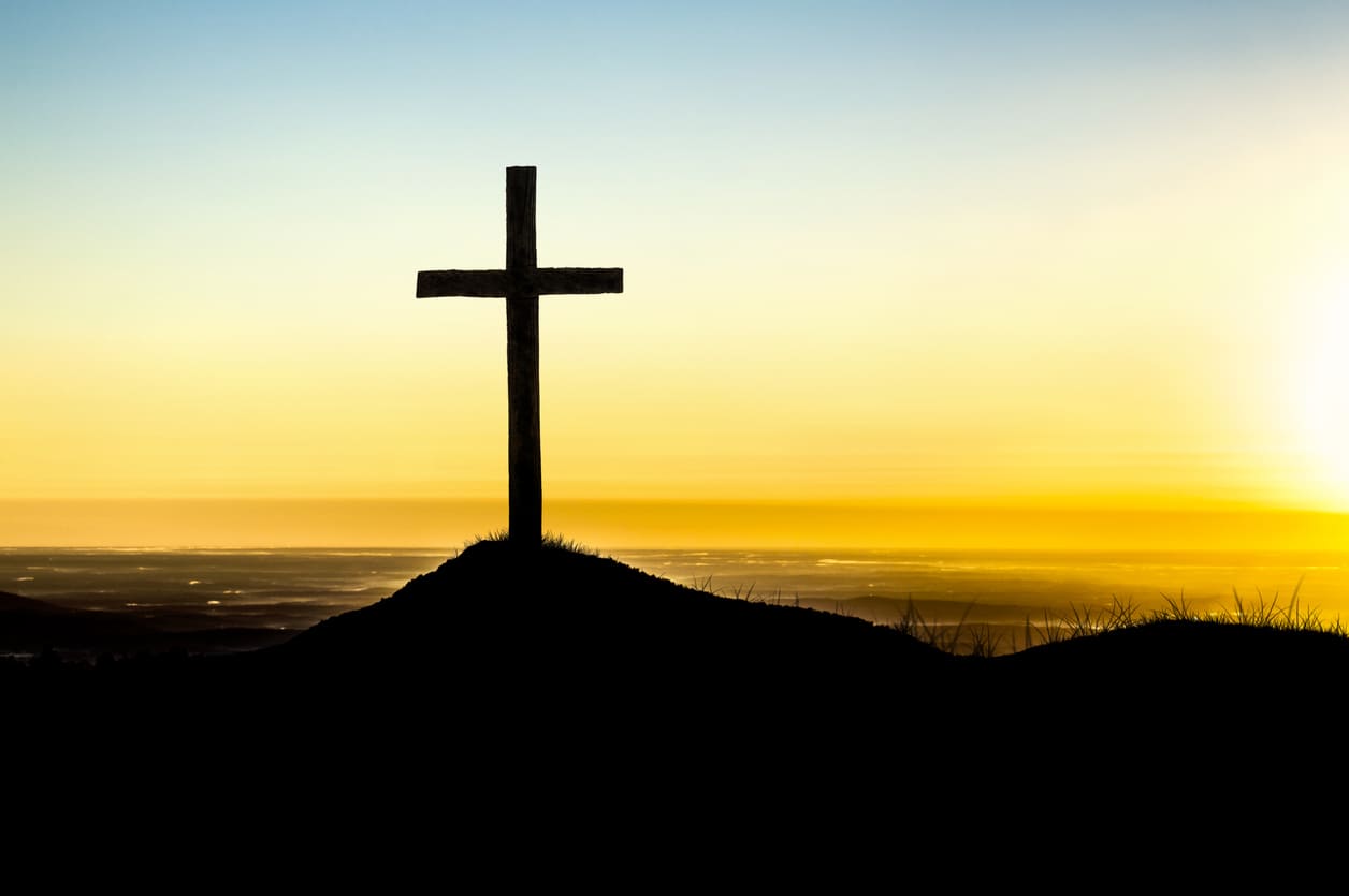 A solitary cross stands on a hill, silhouetted against the golden hues of a peaceful sunrise.