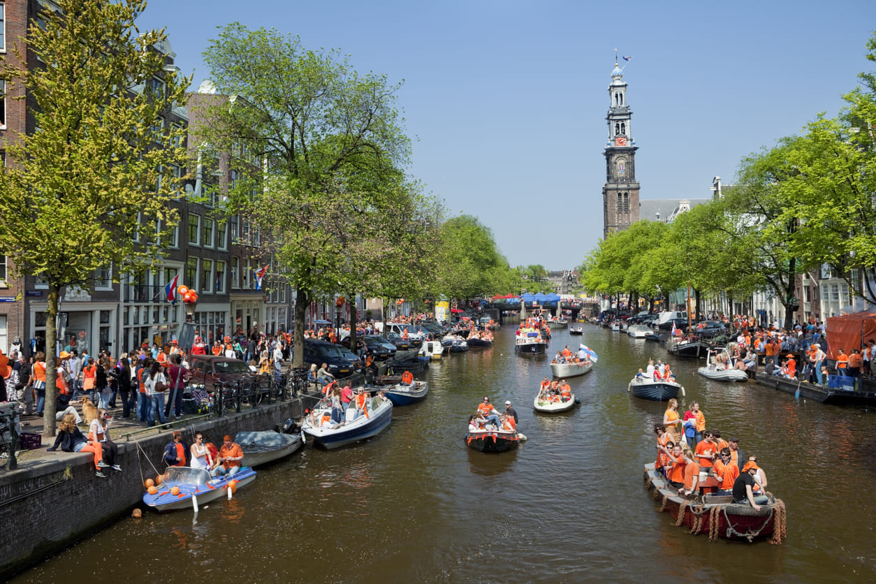 A lively scene of Amsterdam's canals filled with orange-clad revelers on boats, celebrating King's Day under a sunny sky.