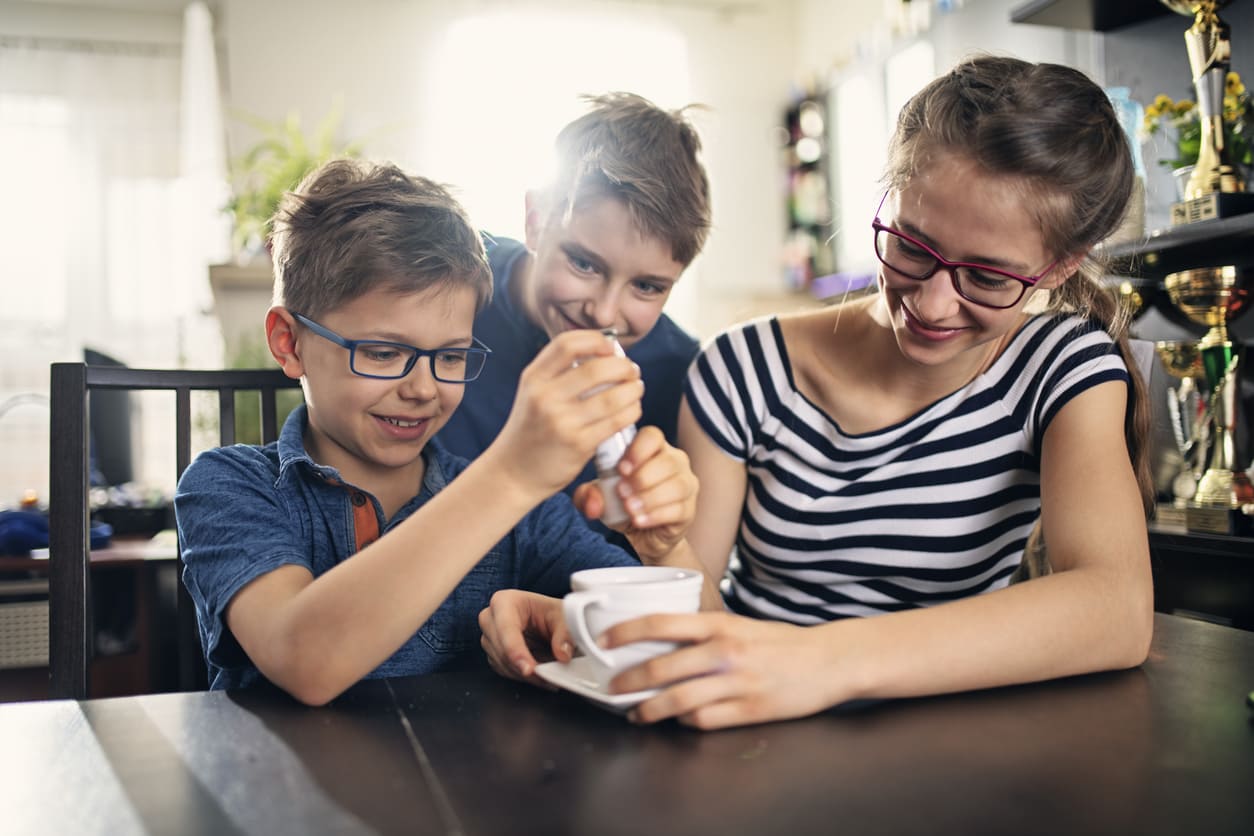 Three smiling children gathered around a table, playfully adding salt to a cup of tea as part of an April Fools' Day prank in New Zealand.
