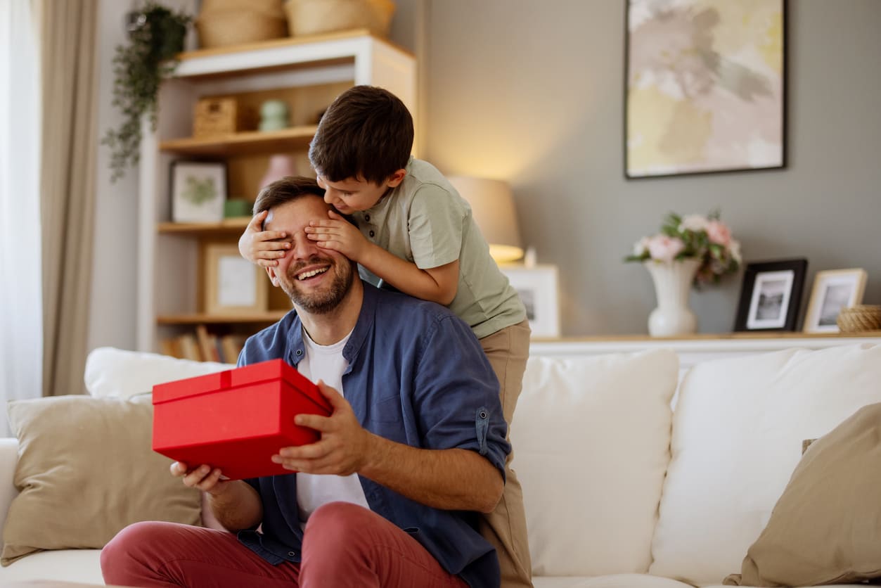 A father and son share a playful moment as the child surprises his dad with a bright red gift box on Father's Day.