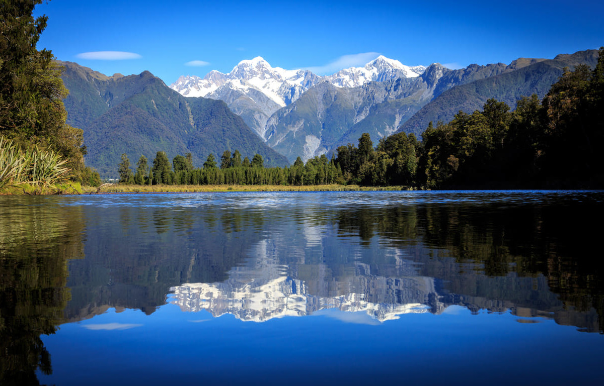 A crystal-clear view of Lake Matheson mirroring the snow-capped Southern Alps, surrounded by lush West Coast rainforest.