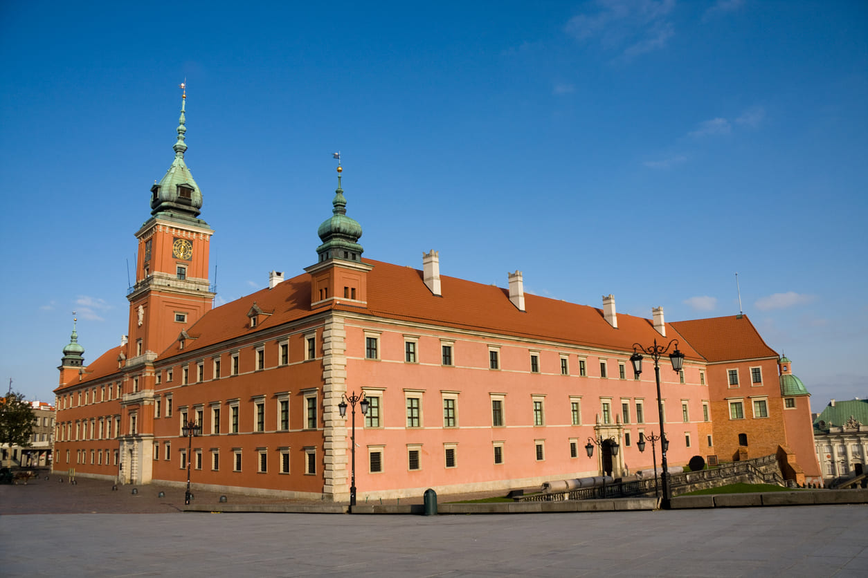 The Royal Castle in Warsaw, a majestic red-brick building with green domed towers under a clear blue sky.