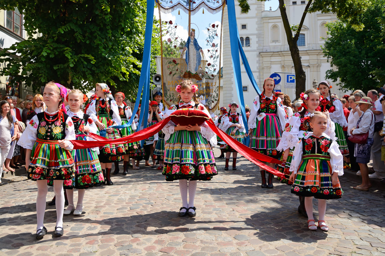 Girls in traditional Polish folk costumes join a Corpus Christi procession, carrying religious banners.