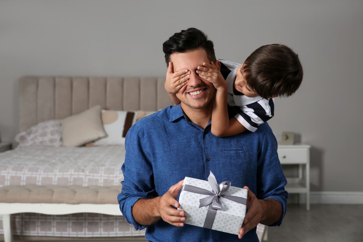 A cheerful father holds a gift while his smiling child playfully covers his eyes, capturing a heartfelt Father's Day moment.