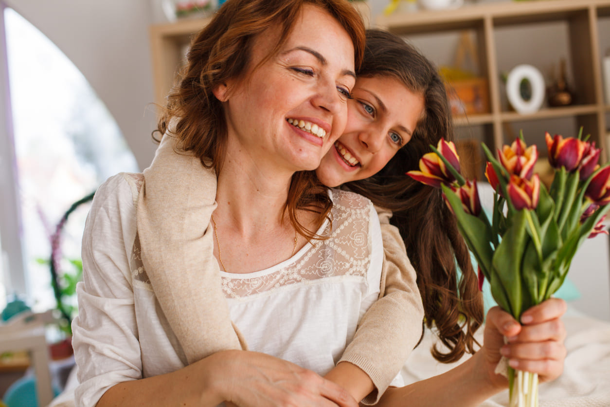 A joyful mother and daughter share a warm embrace, with the mother holding a bouquet of vibrant tulips.