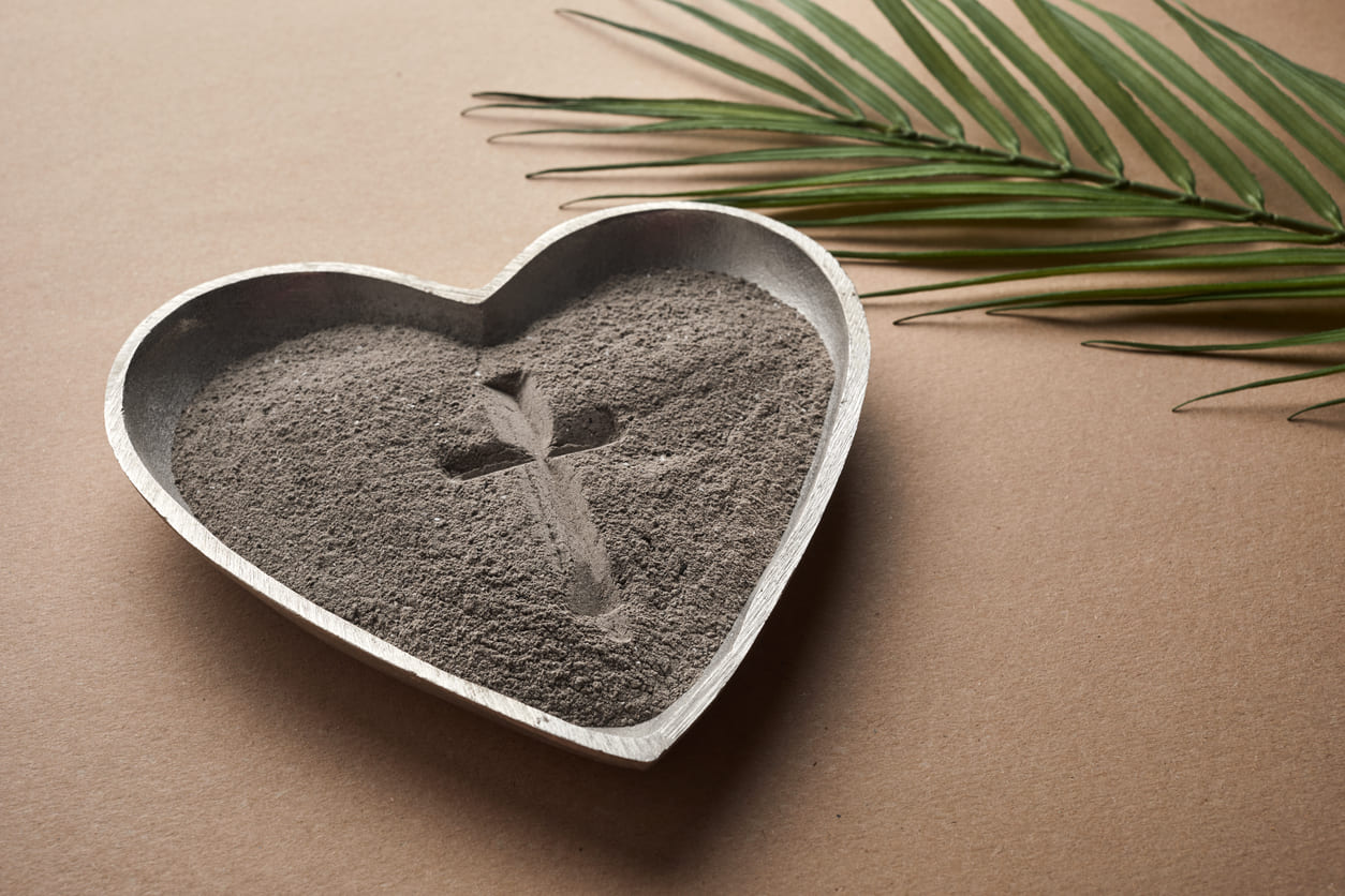 A heart-shaped metal tray filled with ashes, featuring a cross symbol, placed beside a palm leaf on a brown surface, representing Ash Wednesday and the start of Lent.