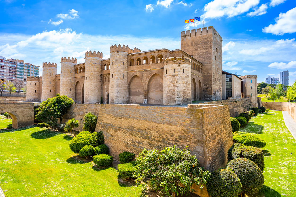 The Aljafería Palace in Zaragoza, a magnificent medieval fortress with Moorish and Gothic influences, stands surrounded by lush greenery under a bright blue sky, symbolizing Aragon's rich heritage.