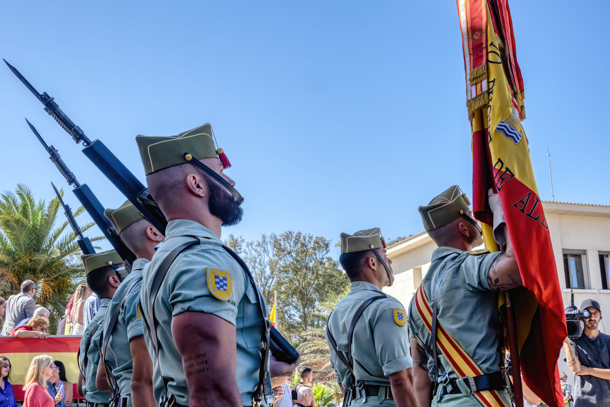 Spanish Legion soldiers in uniform, standing in formation with rifles and a Spanish flag, participating in a ceremonial event on the Day of Ceuta.