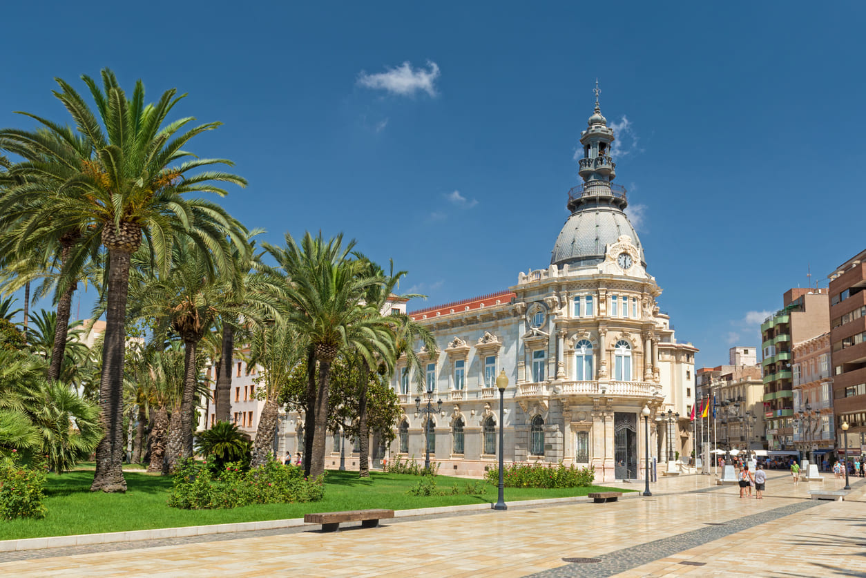 The Palacio Consistorial in Cartagena, Murcia, a stunning Beaux-Arts building, stands elegantly amid lush palm trees under a bright blue sky.