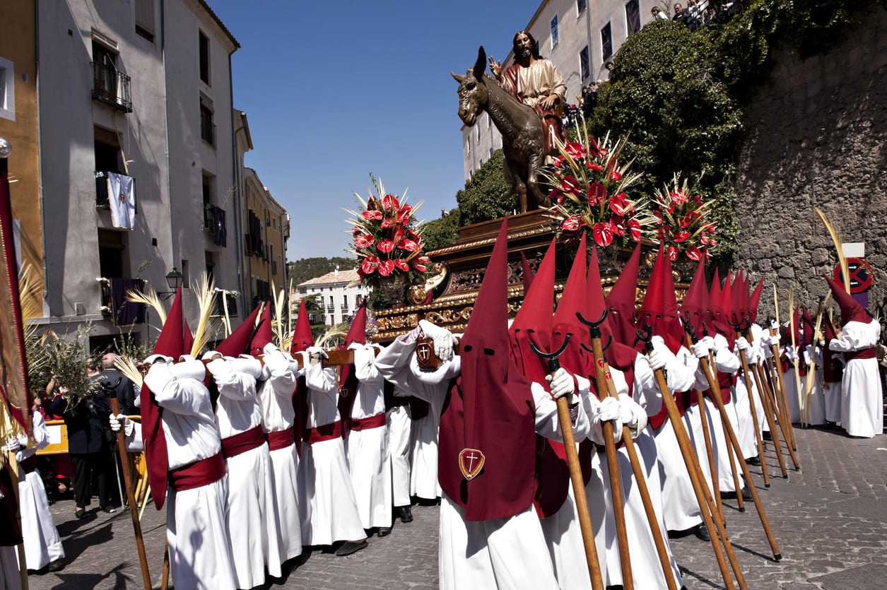 Hooded penitents in white and red robes solemnly carry a paso of Christ, adorned with flowers, during a vibrant Easter Sunday procession in Spain.