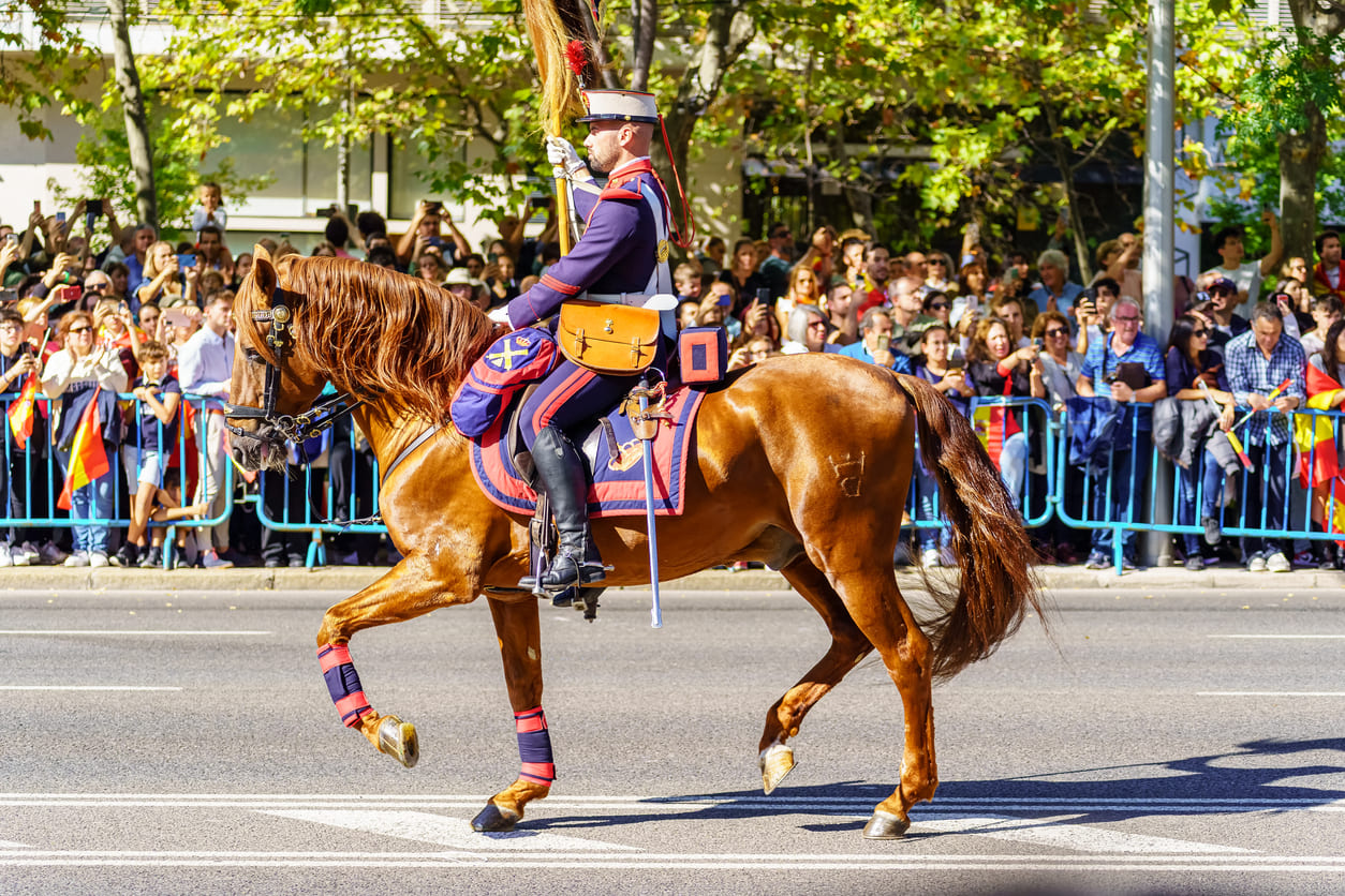 A Spanish soldier on horseback leads a military parade in Madrid, surrounded by crowds waving flags in celebration of Hispanic Day.