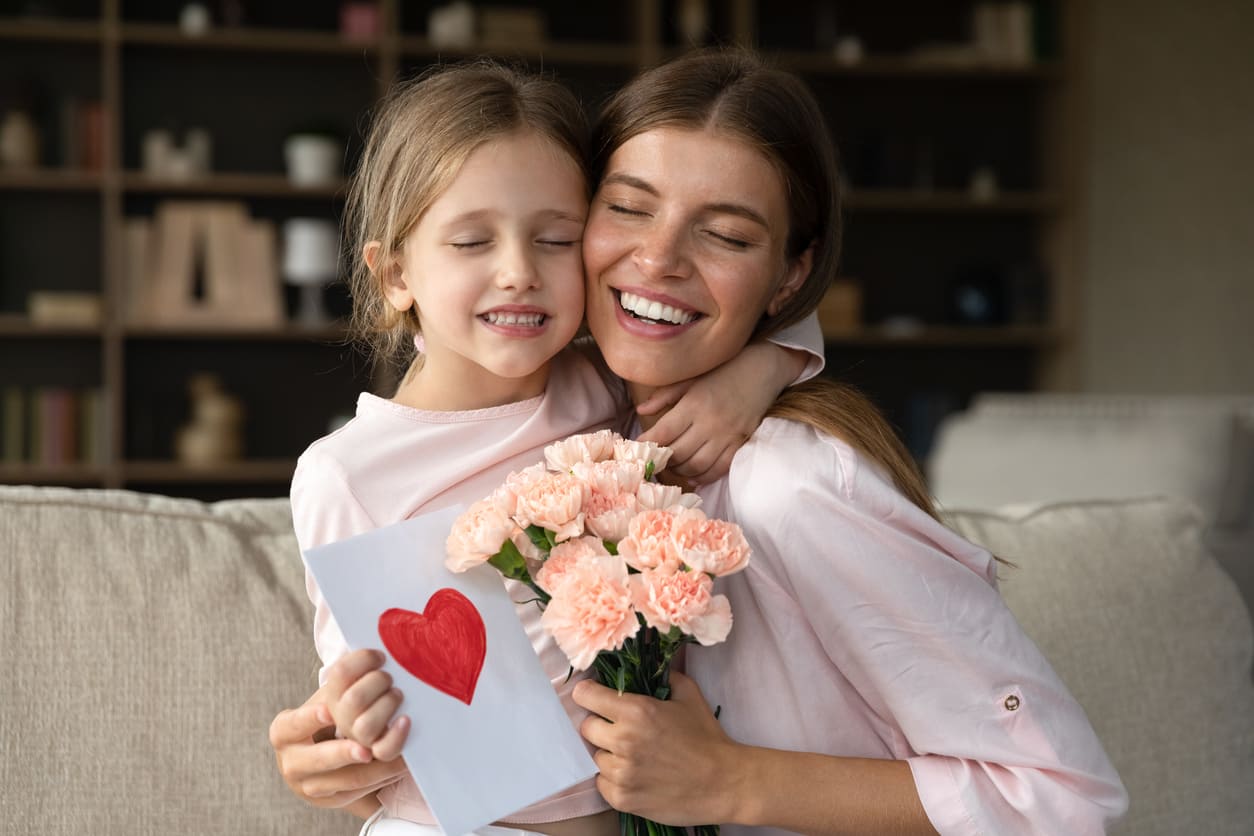 A joyful mother and daughter embrace, holding pink flowers and a handmade card with a red heart.