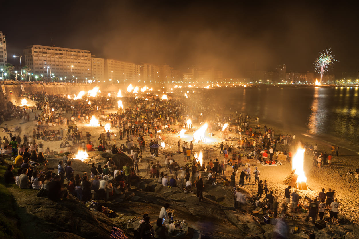 A vibrant beach scene at night with large bonfires, fireworks, and crowds celebrating Saint John the Baptist Day in a lively and festive atmosphere.