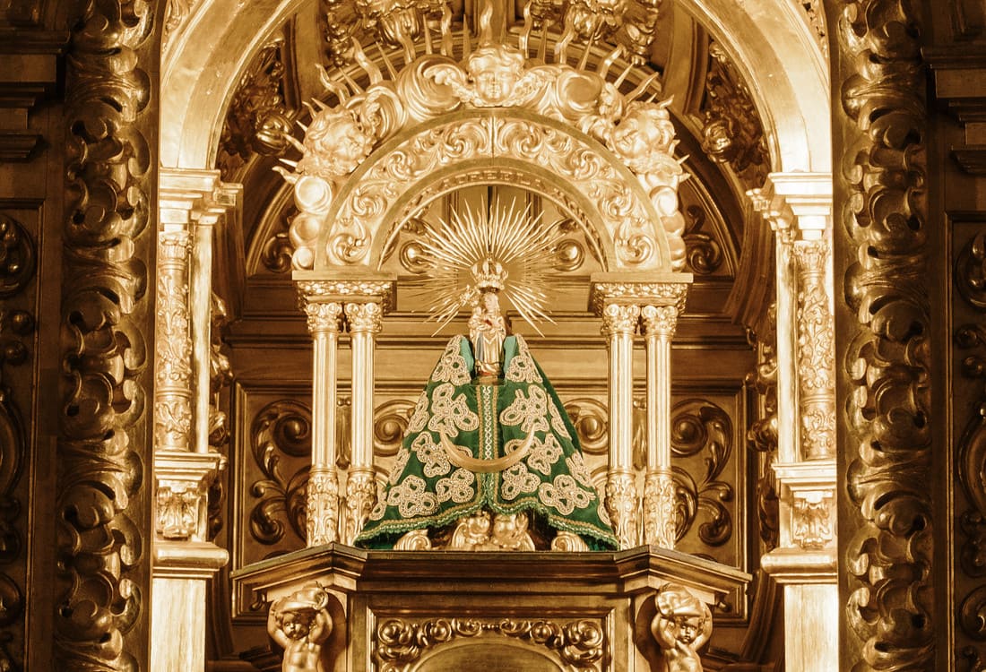 The Virgin of the Bien Aparecida in an opulent golden altar, adorned with a green embroidered mantle and a radiant halo, reflecting deep religious devotion.