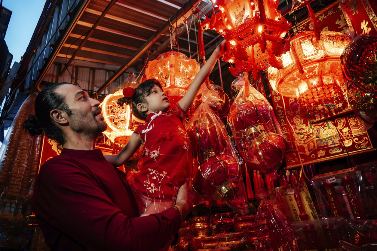 A father and daughter browse a market filled with bright red lanterns and decorations, preparing for Chinese New Year's celebrations.