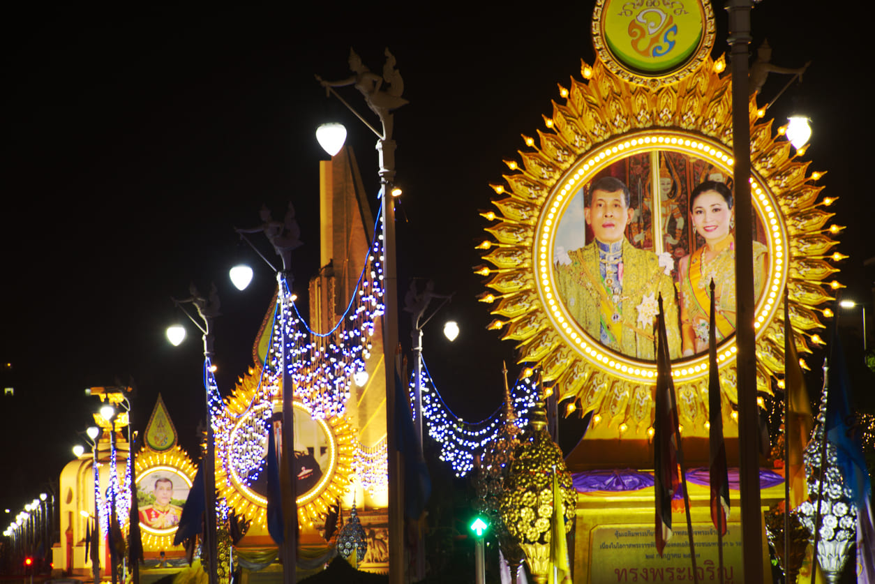 A vibrant display of illuminated royal portraits and decorative lights lining the streets, capturing the grandeur of King Vajiralongkorn's Birthday celebrations.