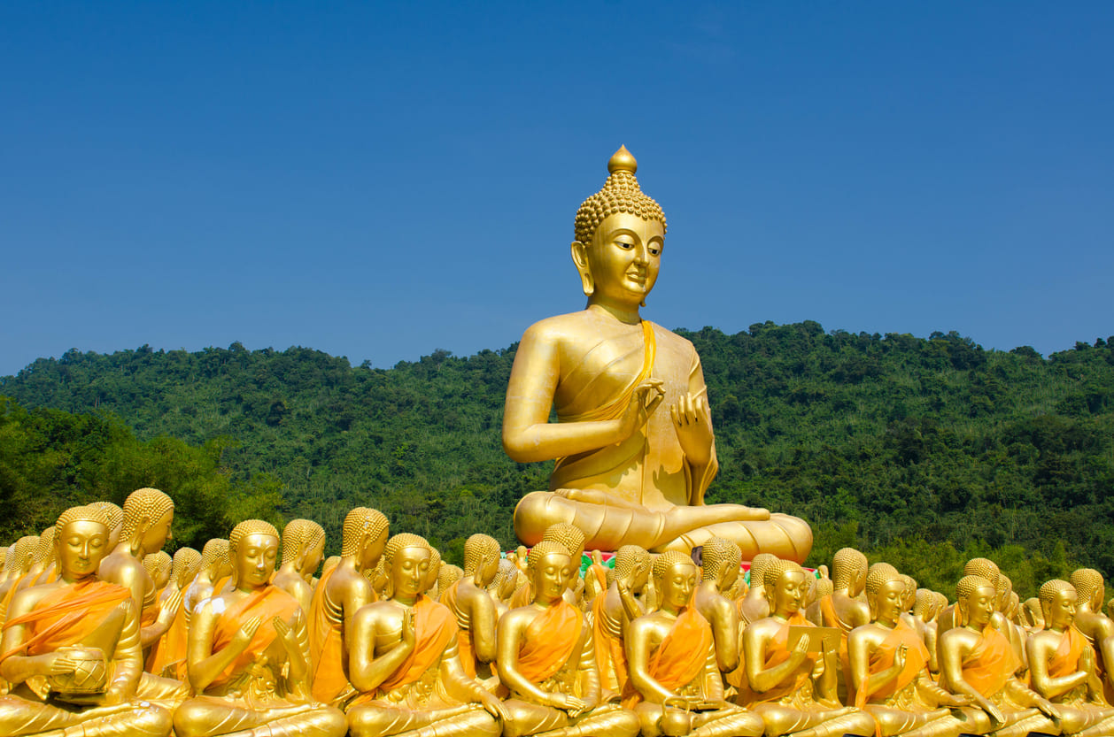 Rows of golden monk statues sit before a large Buddha image, set against lush green hills and a clear blue sky.