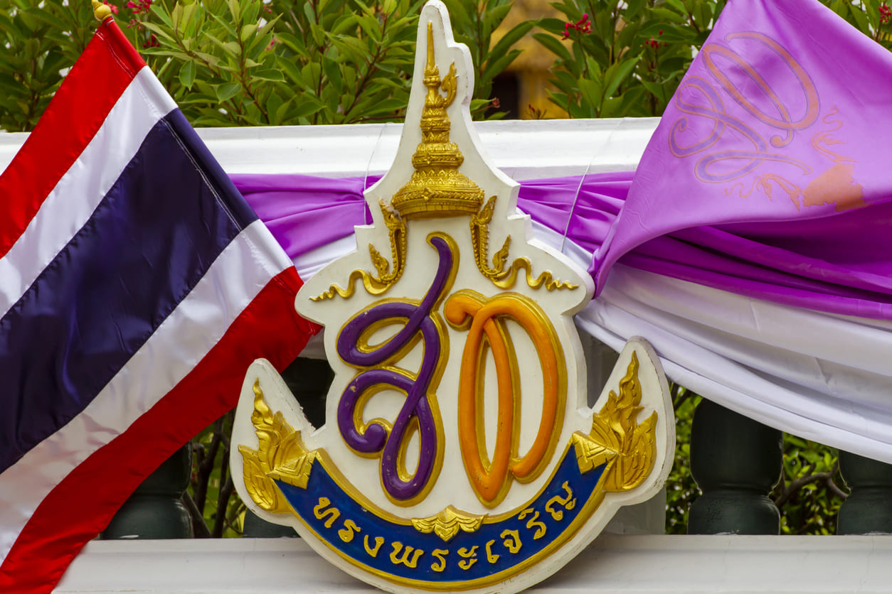 A royal emblem of Queen Suthida framed by purple fabric and the Thai flag, capturing the symbols of devotion displayed during her birthday celebrations.