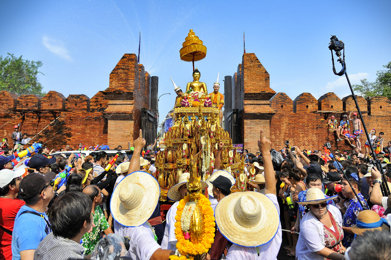A lively crowd sprays water as a golden Buddha statue is paraded through ancient city gates under a clear blue sky.