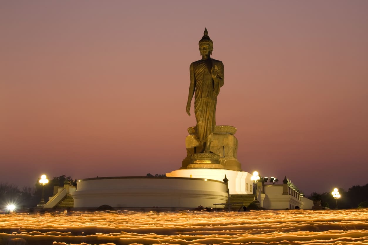 A large standing Buddha statue rises against the evening sky as glowing candlelight trails encircle the temple grounds during the Visakha Bucha procession.