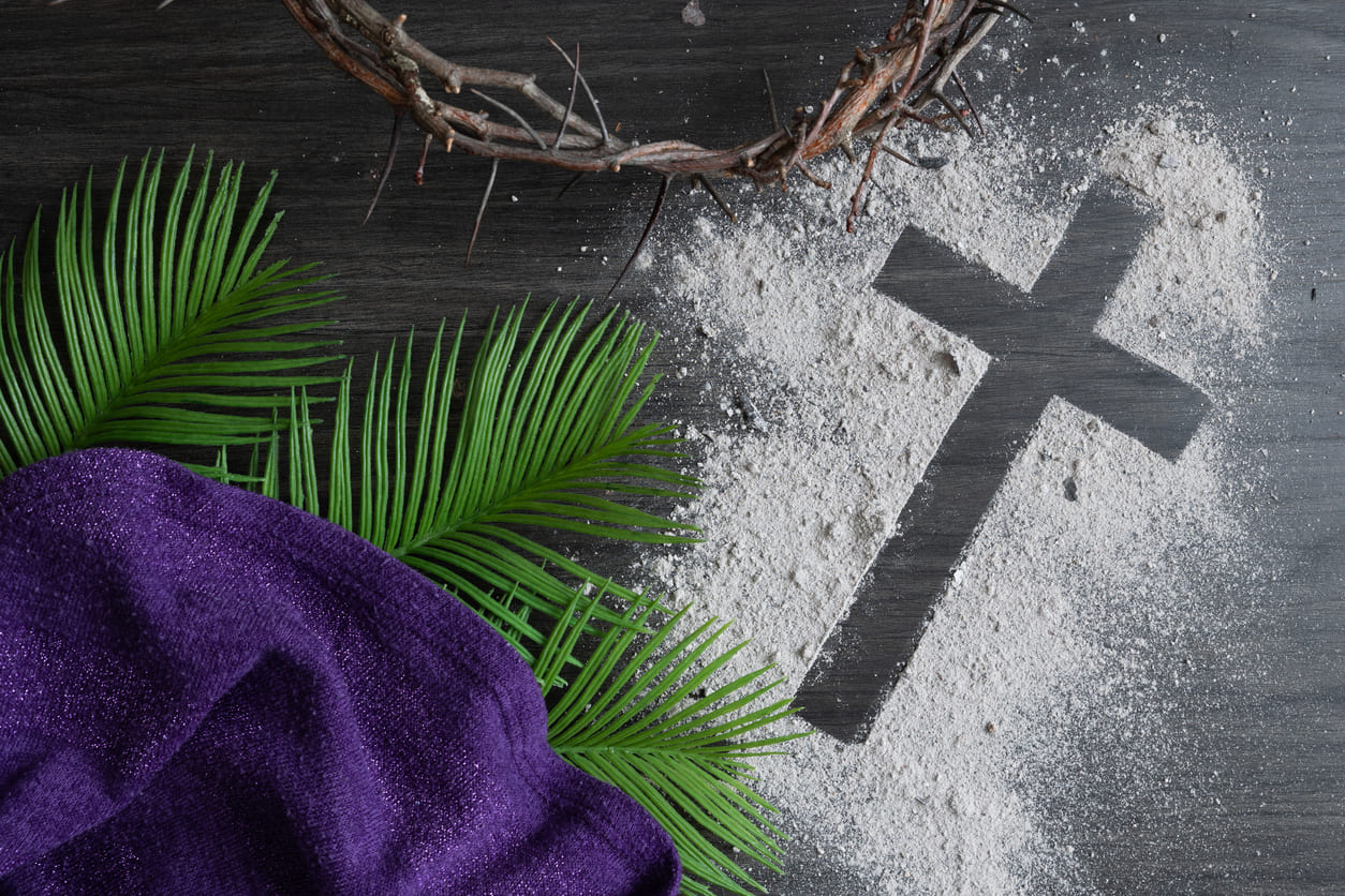 A cross made of ashes lies beside palm leaves, a crown of thorns, and a purple cloth, symbolizing Ash Wednesday and the beginning of Lent.