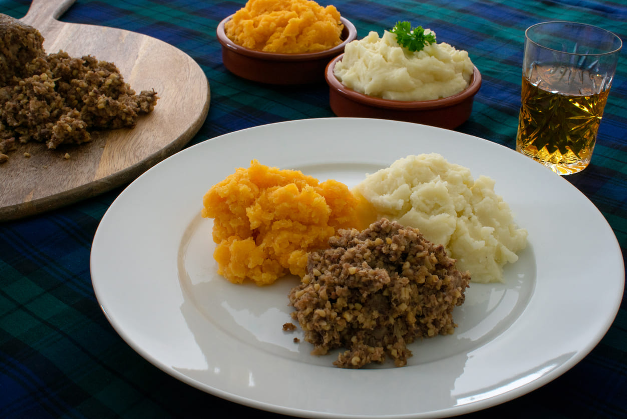 A plate of haggis, neeps, and tatties is served with a glass of whisky, set on a traditional tartan cloth.