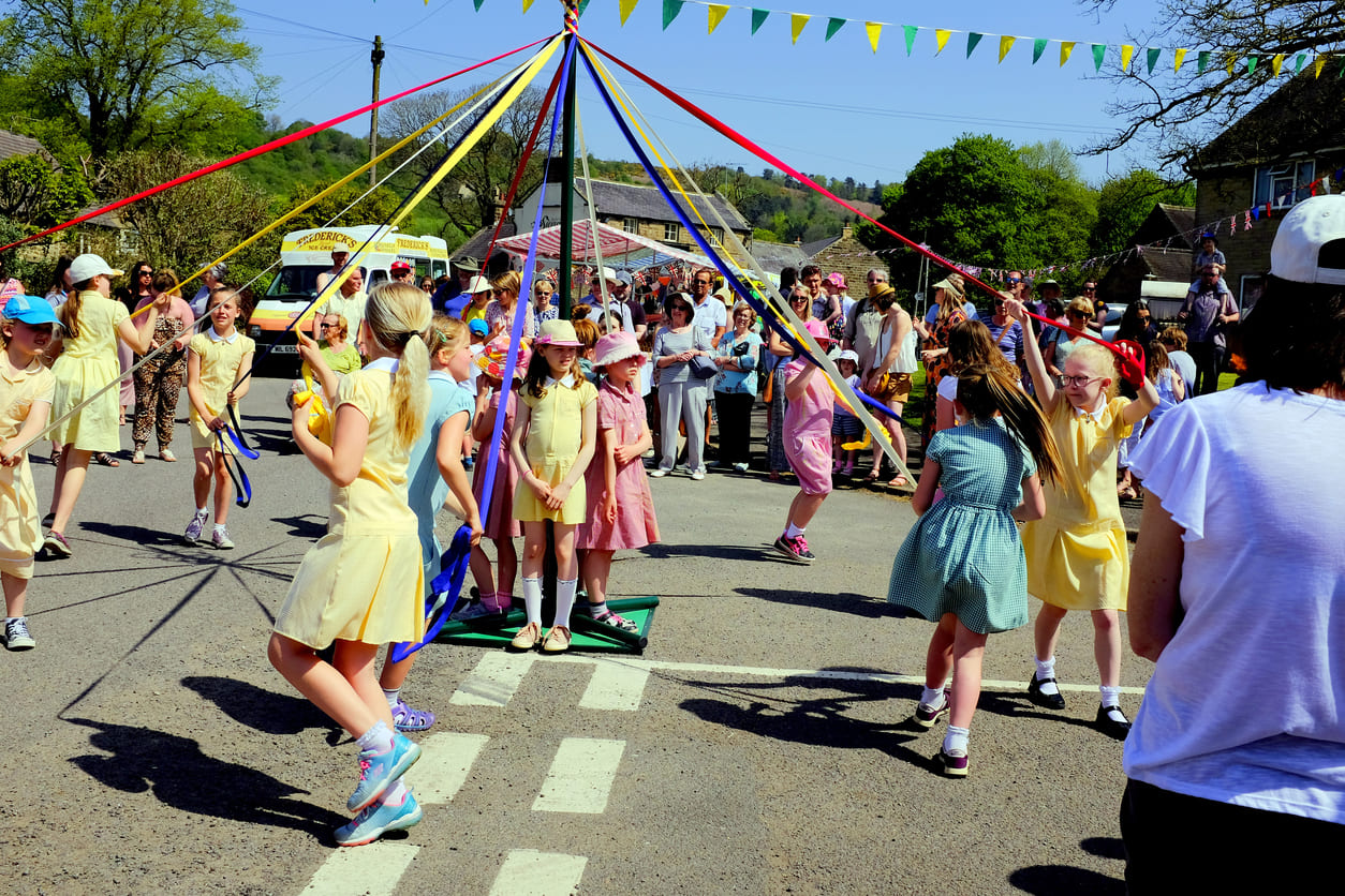 Children joyfully dance around a Maypole on a sunny day, celebrating the Early May Bank Holiday with colorful ribbons and lively festivities.