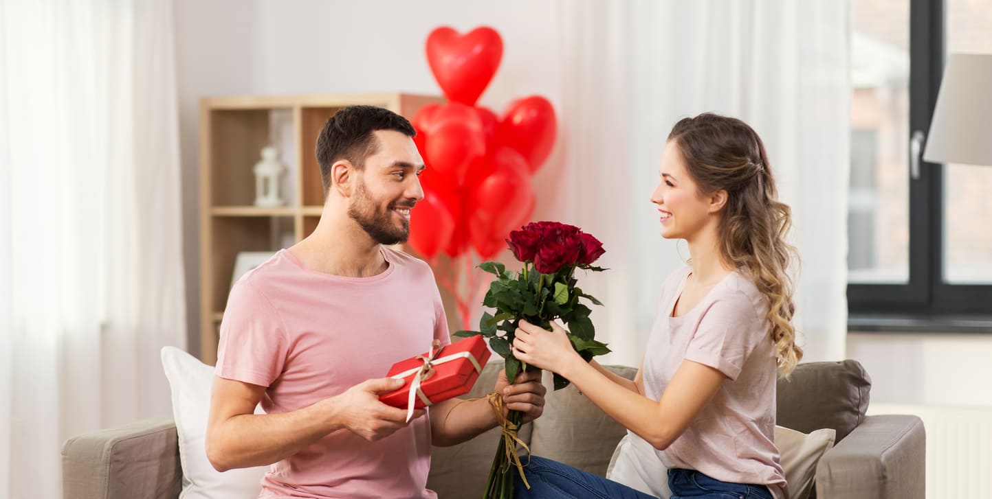 A couple joyfully shares red roses and gifts, surrounded by heart-shaped balloons, celebrating Valentine's Day with love and affection.