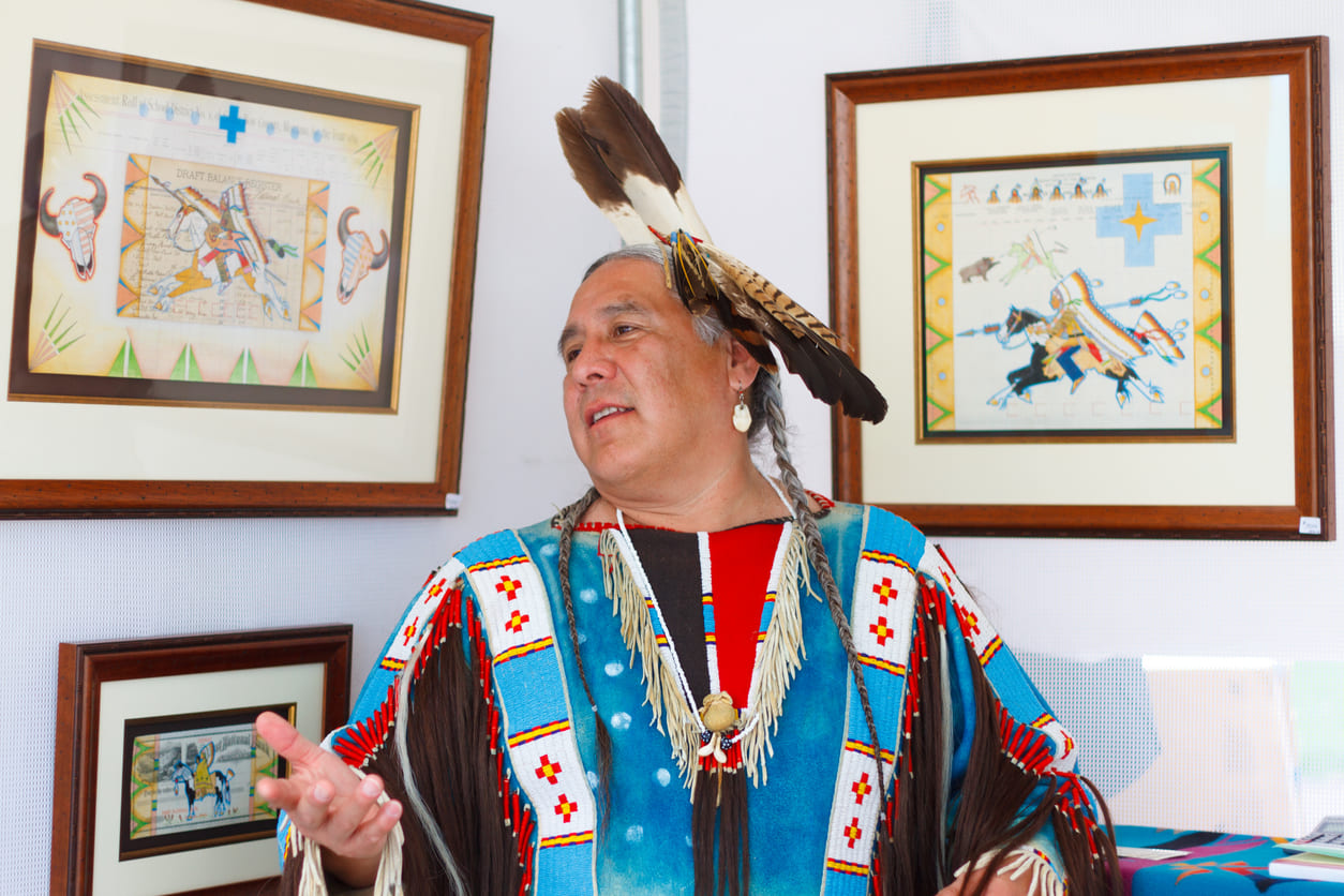 An artist dressed in traditional Native American attire stands beside framed Indigenous artwork, sharing cultural heritage.