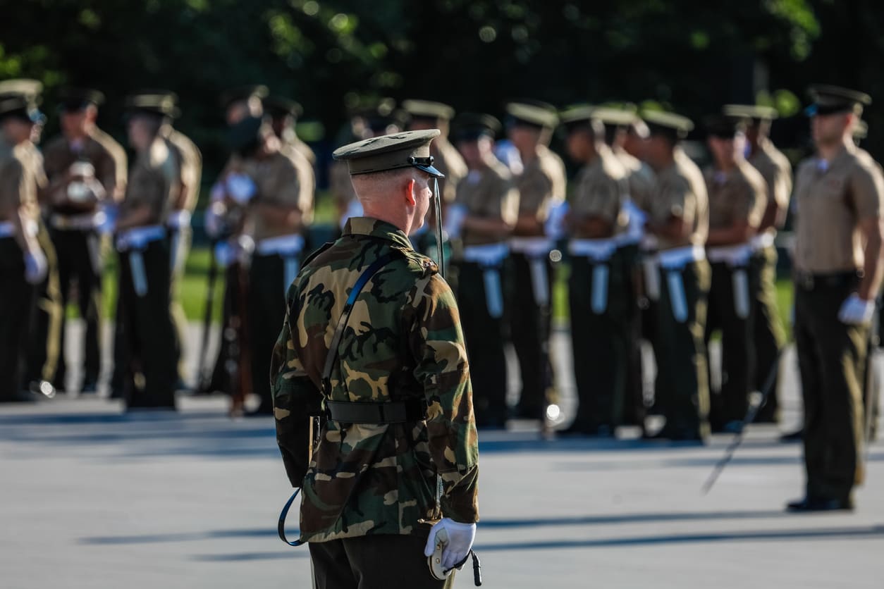 US Army officer in uniform standing in focus, with a group of soldiers assembled in the background during a ceremonial event.