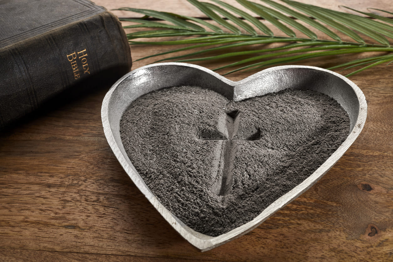 A heart-shaped tray of ash with a cross, alongside a Bible and palm leaves.