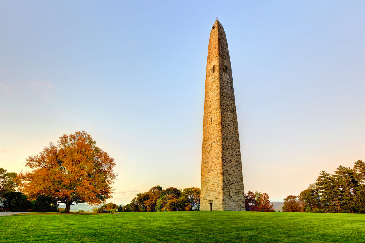 The Bennington Battle Monument, a tall stone obelisk, stands amidst vibrant autumn trees and lush greenery under a clear sky.