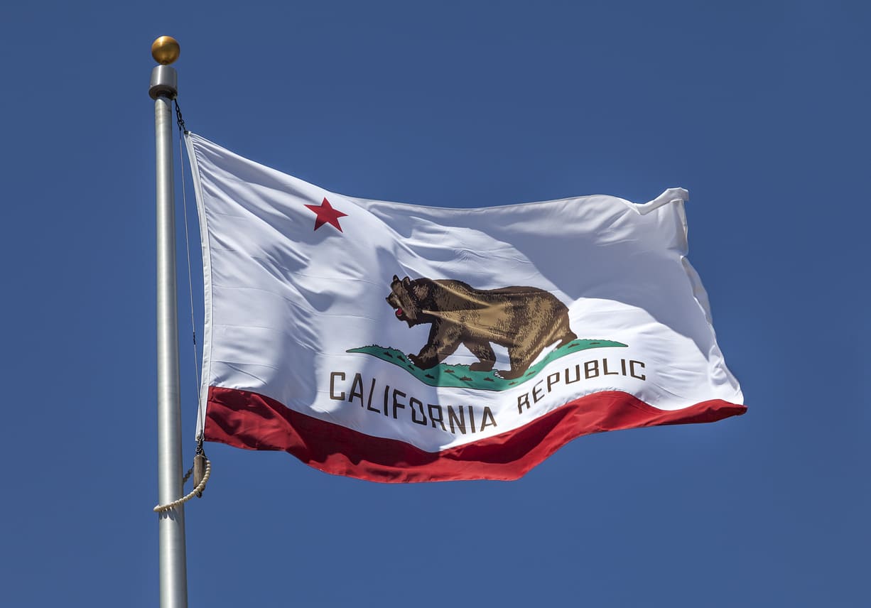 The California state flag with its iconic grizzly bear flies high against a bright blue sky, representing the state's spirit and heritage.