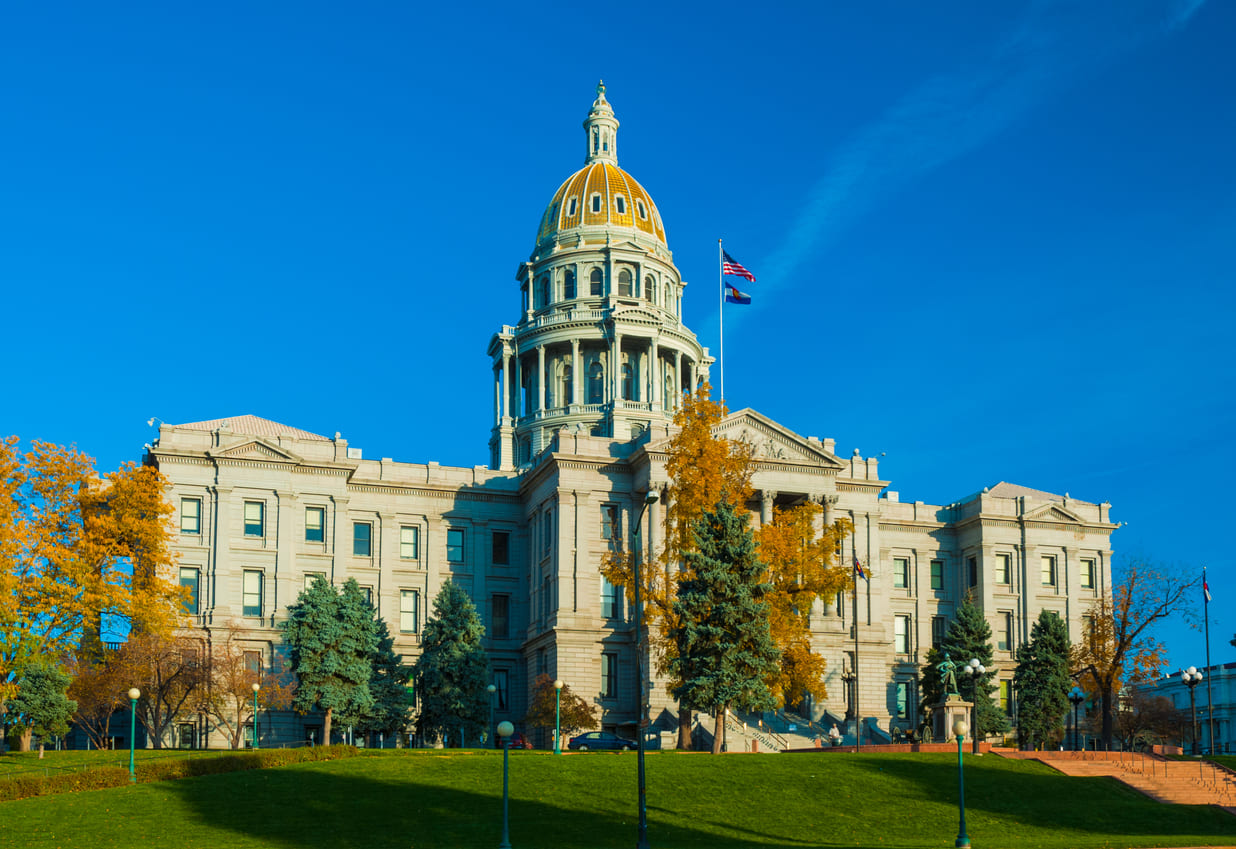 Colorado State Capitol building, a majestic structure with a golden dome, surrounded by vibrant trees under a clear blue sky.