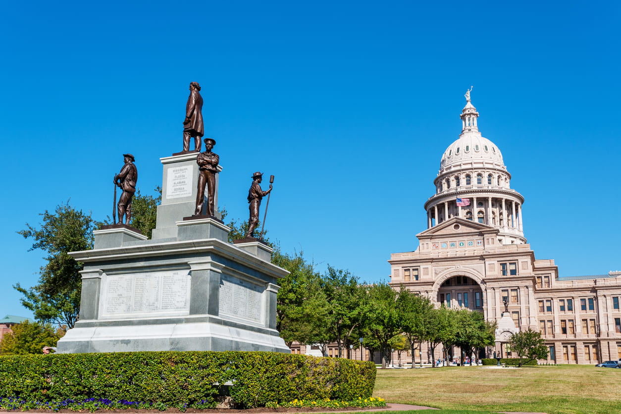 Confederate monument with statues of soldiers near the Texas State Capitol building under a clear blue sky.