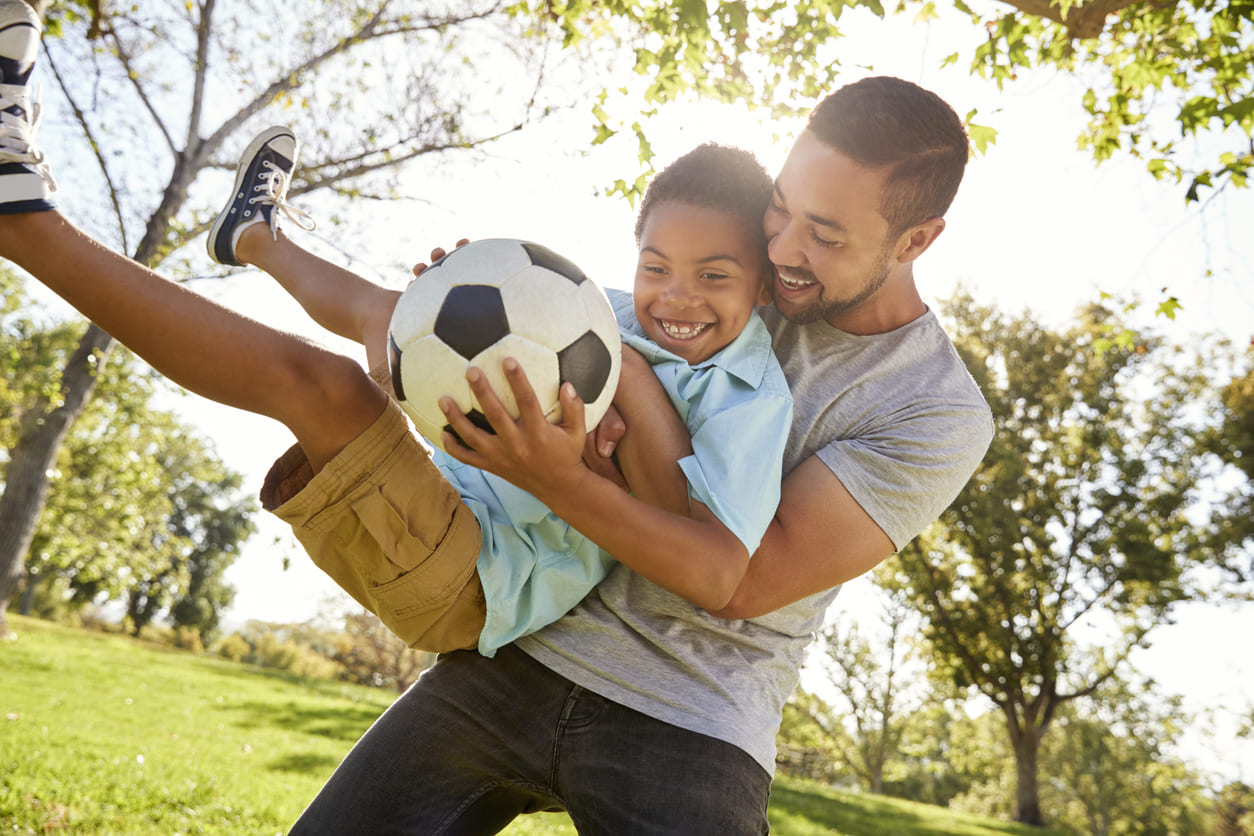 A joyful father and son share a playful moment outdoors with a soccer ball.