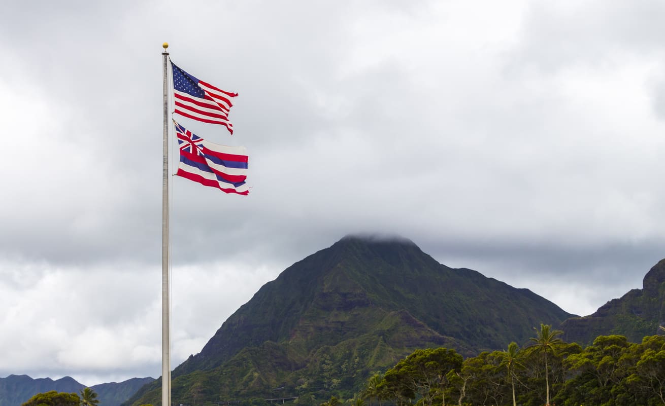 The U.S. and Hawaiian flags wave together against a dramatic mountain backdrop, symbolizing unity and Hawaii's rich cultural heritage.