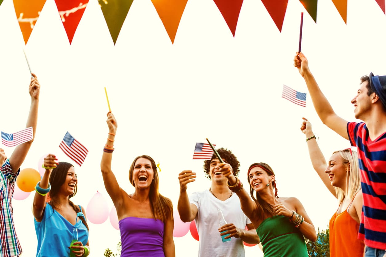 A lively group of young people celebrates together outdoors, waving small American flags and enjoying a festive atmosphere.