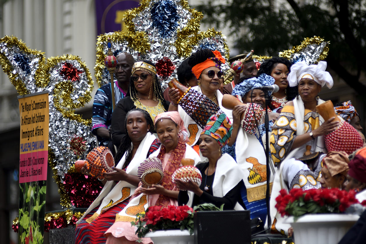 A group of people in colorful traditional attire participate in a Juneteenth parade, celebrating African and Caribbean heritage with music and festive decorations.